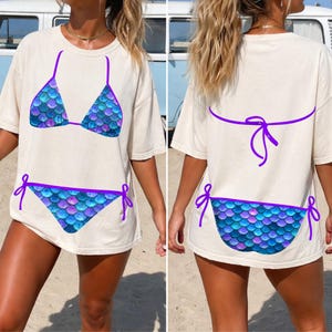 May include: A beige short-sleeve t-shirt featuring a graphic print of a bikini with a blue and purple mermaid scale pattern. The front displays the bikini top and bottom, while the back shows the bikini bottom and the tied straps of the top.
