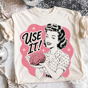 May include: A cream-colored t-shirt featuring a graphic of a person with dark hair holding a pink brain. The text "USE IT!" is printed in black next to the person. The design is set against a pink background with small white star accents.