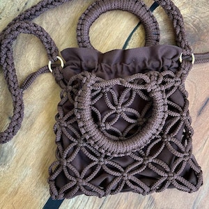 以下が含まれることがあります： 茶色のマクラメバッグ。編み込みのハンドルと、お揃いのショルダーストラップが付いています。バッグの前面には円形のデザインがあり、巾着開閉式です。バッグは柔らかい織り素材で作られています。