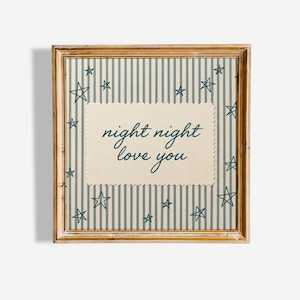 May include: A square wooden frame containing a print with a light blue and white vertical striped background featuring dark blue hand-drawn stars. A central cream-colored rectangular patch with scalloped edges displays the text night night love you.