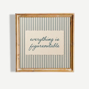 Può includere: Un oggetto da parete quadrato in una cornice di legno marrone chiaro con un centro in tessuto color crema dai bordi smerlati. Lo sfondo presenta strisce verticali blu e crema. Il testo "everything is figureoutable" è stampato in blu scuro.