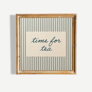 May include: A square wall art piece in a light brown wooden frame featuring a background of vertical sage green and cream stripes. Centered is a cream-coloured rectangular card with scalloped edges and the dark green cursive text time for tea.