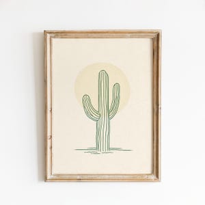 Op de afbeelding: Een ingelijste print met een minimalistische illustratie van een groene cactus tegen een lichtgele cirkel op een beige achtergrond. Het kunstwerk zit in een lichtbruine houten lijst, geschikt voor woondecoratie.