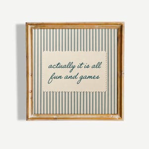 May include: A square wall art piece in a light brown wooden frame featuring a background of vertical grey and white stripes. Centered is a cream-colored rectangular fabric patch with scalloped edges displaying the text "actually it is all fun and games" in dark green cursive script.