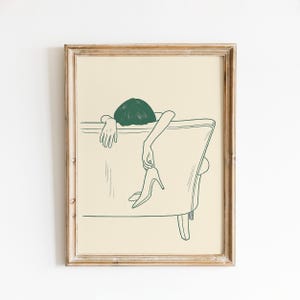 Peut inclure: Impression artistique encadrée présentant un dessin au trait minimaliste. L'œuvre représente une personne se reposant sur une chaise, avec une coupe au carré verte et tenant un escarpin. La palette de couleurs est un vert sourd sur fond crème.