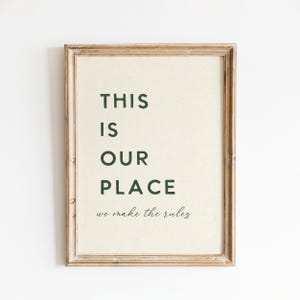 May include: A framed print with the words "THIS IS OUR PLACE" in dark green, and the phrase "we make the rules" in a script font. The frame is a light brown colour.