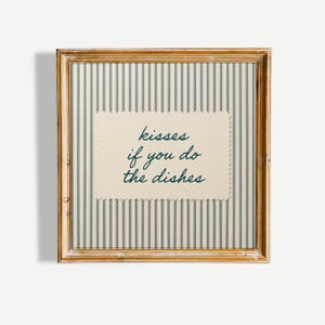 May include: A square wall art piece in a light brown wooden frame featuring a beige background with vertical dark grey stripes. Centered is a cream-colored rectangular card with scalloped edges and the dark green text: kisses if you do the dishes.