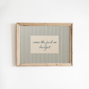 May include: A rectangular wooden frame containing a print with a vertical blue and beige striped background. Centered is a beige scalloped-edge rectangular card featuring the black cursive text "come the fuck on bridget" printed in the middle.