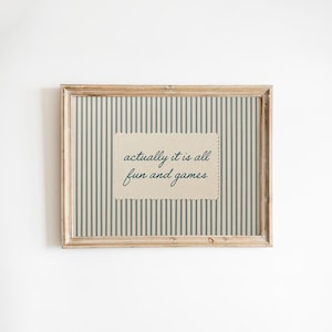 May include: A rectangular wooden frame containing a print with a vertical blue and beige striped pattern. Centered on the print is a rectangular beige card with scalloped edges featuring the dark blue cursive text, actually it is all fun and games.