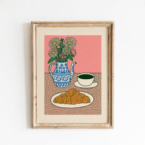 May include: A framed art print featuring a blue and white patterned teapot filled with white daisies, a white teacup with dark green liquid, and a golden-brown croissant on a white plate, all set against a pink background with green dots.
