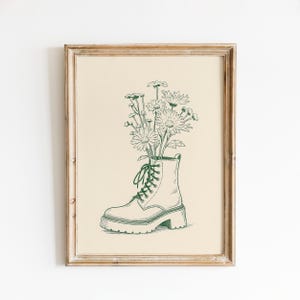 May include: A framed art print featuring a green line drawing of a combat boot filled with daisies. The boot has a chunky sole and is laced up. The daisies have multiple petals and tall stems. The background is a light beige color.