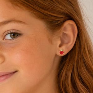 Puede incluir: Un pequeño pendiente de botón con forma de manzana roja y un detalle de hoja verde. La manzana está delineada en metal dorado y presenta un relleno de esmalte rojo vibrante. El pendiente se lleva en el lóbulo, mostrando un diseño compacto.
