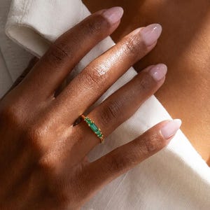 Puede incluir: Un anillo de oro con una piedra preciosa verde central en talla baguette, flanqueada por dos piedras verdes más pequeñas en talla redonda a cada lado. La banda es fina y metálica, con un acabado en oro pulido que resalta las piedras.