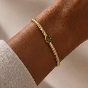 Pode incluir: Uma pulseira de tom dourado com um design de banda texturizada e canelada. O centro da pulseira é adornado com uma pequena pedra preciosa verde de formato oval, engastada num bisel liso de tom dourado, criando uma peça de joalharia simples.