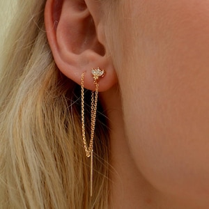 May include: A close-up of a gold earring with a small, sparkling floral design. The earring features a delicate chain that dangles down. The earring is worn on the ear of a person with blonde hair.