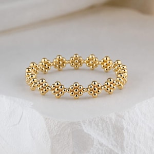 May include: A gold ring featuring a continuous band composed of small, spherical beads arranged in repeating flower-like clusters. The metallic surface has a polished finish, creating a series of interconnected, rounded shapes that form the circular structure.