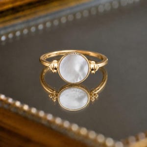 Peut inclure: Une bague en or dotée d'une incrustation circulaire en nacre blanche irisée au centre. L'anneau est fin et poli, avec de petits accents métalliques décoratifs entourant le sertissage circulaire des deux côtés, créant un design équilibré.