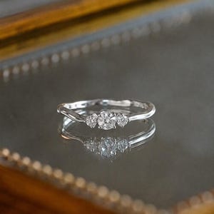 Peut inclure: Une bague argentée dotée d'un anneau ondulé serti de trois pierres rondes et transparentes. La pierre centrale est légèrement plus grande que les deux pierres latérales, toutes maintenues par des griffes métalliques, formant un design symétrique.