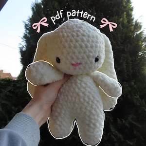 Puede incluir: Un peluche de conejo de ganchillo hecho a mano, de color crema, con largas orejas caídas, ojos negros y una nariz rosa. El texto "pdf pattern" está escrito encima del conejo. El conejo se sostiene frente a un fondo verde y frondoso.