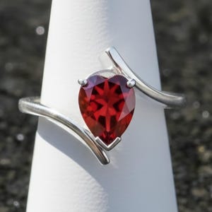 Op de afbeelding: Een zilveren ring met een peervormige, dieprode edelsteen. De band van de ring buigt om de steen heen, wat een modern, elegant ontwerp creëert. De ring wordt getoond op een witte, conische standaard tegen een donkere, gespikkelde achtergrond.