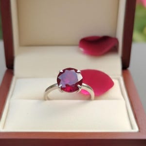 Op de afbeelding: Een zilveren ring met een grote, ronde, dieprode edelsteen ligt in een houten sieradendoosje met een crèmekleurig interieur. Rozenblaadjes omringen de doos, met een vage bloemenachtergrond.