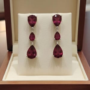 Op de afbeelding: Een paar elegante oorhangers met drie dieprode, peervormige edelstenen, gezet in zilver. De oorbellen worden gepresenteerd in een open houten sieradendoos met een crèmekleurig interieur, wat het luxueuze ontwerp van de sieraden benadrukt.