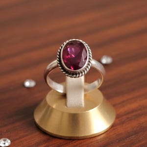 Op de afbeelding: Een zilveren ring met een grote, ovale, dieproze edelsteen. De edelsteen is gezet in een gedraaide, touwachtige rand. De ring wordt getoond op een goudkleurige standaard tegen een donkere houten achtergrond. Verschillende kleine, heldere kristallen zijn verspreid.
