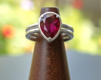 Puede incluir: Un anillo de plata con una piedra preciosa roja oscura en forma de pera. El anillo se exhibe en un soporte de madera. El fondo está borroso, con follaje verde y amarillo.