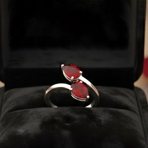 Op de afbeelding: Een zilveren ring met twee peervormige, rode edelstenen, gepresenteerd in een open zwarte fluwelen ringdoos. Zachte verlichting en rozenblaadjes op de achtergrond suggereren een romantisch thema.