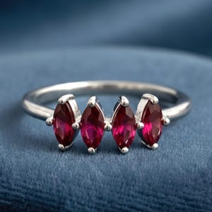 Op de afbeelding: Een zilveren ring met vier marquise-geslepen, dieprode edelstenen. De edelstenen zijn in een rij over de bovenkant van de ring geplaatst. De ring wordt gepresenteerd op een blauw fluwelen oppervlak.