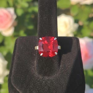 Op de afbeelding: Een ring met een grote, vierkant geslepen, rode edelsteen, gepresenteerd op een zwarte fluwelen standaard. De ring heeft een zilveren band met kleine accentstenen. De achtergrond toont wazige roze en witte rozen.