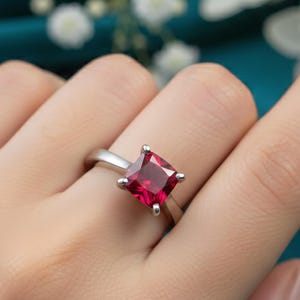 Op de afbeelding: Een zilveren ring met een vierkante, helderrode edelsteen. De ring wordt aan een vinger gedragen, waarbij de kleur van de edelsteen en het eenvoudige ontwerp van de ring worden benadrukt. De achtergrond is turquoise met witte bloemen.