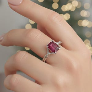 Op de afbeelding: Een zilveren ring met een grote, rechthoekige, dieproze edelsteen. De band van de ring heeft een delicaat, verweven ontwerp. De ring wordt getoond op een hand met een manicure in neutrale tinten.