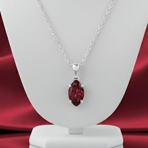 Può includere: Una collana d'argento con un pendente a forma di pietra preziosa rosso scuro taglio marquise. Il pendente è incastonato in una lunetta d'argento e pende da una delicata catena d'argento. La collana è esposta su un busto bianco su sfondo rosso.