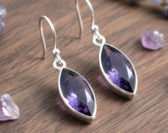 May include: Silver earrings featuring marquise-cut, deep purple amethyst gemstones. The earrings are presented on a wooden surface, alongside small amethyst crystals and lavender sprigs.