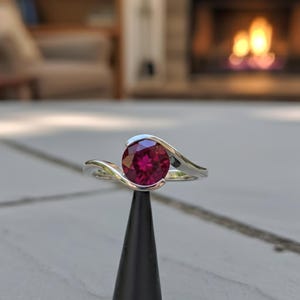 Op de afbeelding: Een zilveren ring met een grote, ronde, dieprode edelsteen. De band van de ring buigt om de steen heen en creëert een modern design. De ring wordt getoond op een zwarte standaard, met een vage achtergrond van een open haard.