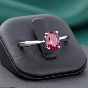 Op de afbeelding: Een zilveren ring met een grote, ovaal geslepen, dieproze edelsteen. De ring wordt gepresenteerd op een zwarte sieradenstandaard tegen een donkergroene achtergrond. De edelsteen is het middelpunt en toont zijn levendige kleur en helderheid.