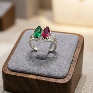 Op de afbeelding: Een zilveren ring met twee marquise-geslepen edelstenen, één smaragdgroen en één robijnrood. De ring heeft een delicaat bloemmotief op de band en wordt gepresenteerd op een grijze fluwelen ondergrond in een houten sieradendoos.