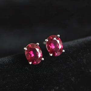 Op de afbeelding: Een paar ovale, robijnrode edelsteen oorknopjes. Elke oorbel heeft een levendige, gepolijste edelsteen, gezet in een zilverkleurige prong setting. De oorbellen worden gepresenteerd op een zwart fluwelen oppervlak.