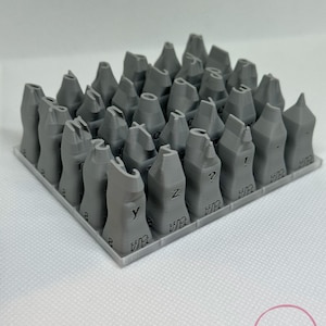 Puede incluir: Un conjunto de sellos de letras de cerámica gris impresos en 3D, dispuestos en una cuadrícula. Cada sello presenta una letra diferente del alfabeto, con las letras "Y" y "Z" visibles. Los sellos están sobre una superficie blanca.