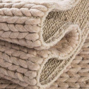May include: Close-up of a beige woven rug with a thick, braided border. The rug's texture is visible, with a combination of tightly woven sections and a raised, rope-like design. The rug is folded over, showing its layered construction.