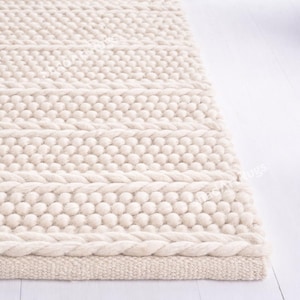 Puede incluir: Alfombra crema con borde trenzado y filas de bolitas. El diseño de la alfombra presenta patrones alternos de bolitas y líneas horizontales, creando una superficie táctil y visualmente atractiva. La alfombra está hecha de un material suave de tono neutro.