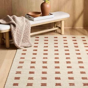 May include: Cream-colored rectangular rug with a repeating pattern of rust-colored rectangular blocks. The rug is placed on a hardwood floor. A bench with a white cushion and a wooden tray with a vase and bowl are in the background.
