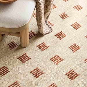 May include: A cream-coloured area rug featuring a repeating pattern of small, raised, rust-coloured rectangular blocks arranged in a grid. The rug has a textured, woven surface and is partially covered by a wooden stool with a beige cushion and a chunky knit throw.