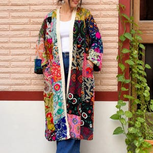 Könnte beinhalten: Lange Jacke im Kimono-Stil mit Patchwork und einer lebendigen Mischung aus Farben und Mustern. Die Jacke hat eine cremefarbene Paspel und wird über einem weißen Oberteil und einer blauen Jeans getragen. Das Design umfasst florale und geometrische Motive.