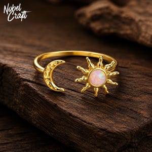 May include: A gold-toned open ring featuring a crescent moon on one end and a sun motif on the other. The sun design is centered with a round, iridescent opal-like stone. The text Nobel Craft is visible in the upper left corner of the image.