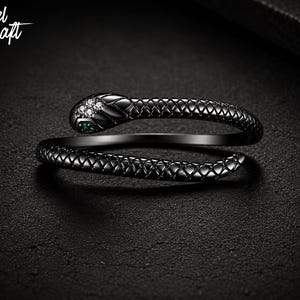 May include: A black metal snake ring featuring a textured scale pattern along the band. The snake head is adorned with four small clear stones and two small green stones for eyes. The text Nobel Craft is visible in the upper left corner.