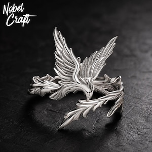 May include: A silver-toned ring featuring a detailed bird with outstretched wings and textured plumage. The band is formed by stylized leaf-like elements that wrap around the finger. The text Nobel Craft is visible in the upper left corner.