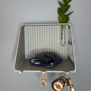 Puede incluir: Estante de pared gris y blanco. Una cartera azul descansa sobre el estante, con un pequeño jarrón de vidrio que contiene hojas verdes. Debajo, un llavero de búho y un llavero con llaves.