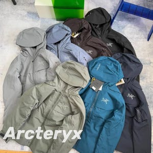 Könnte beinhalten: Eine Auswahl an Kapuzenjacken von Arc'teryx in verschiedenen Farben, darunter Grau, Blau und Schwarz. Jede Jacke hat einen Reißverschluss, eine Kapuze und das Arc'teryx-Logo. Die Jacken sind auf einer hellen Oberfläche angeordnet.